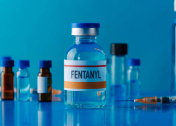 closeup of a simulated vial of fentanyl on a blue table next to a syringe and some other different vials